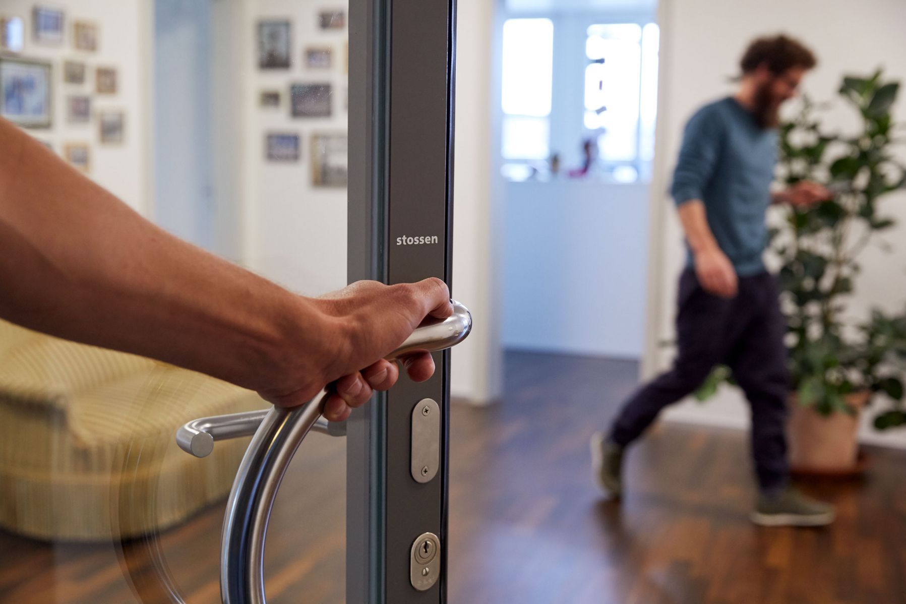 Eine Hand öffnet eine Eingangstür, während im Hintergrund eine weitere Person durch den Eingangsbereich geht