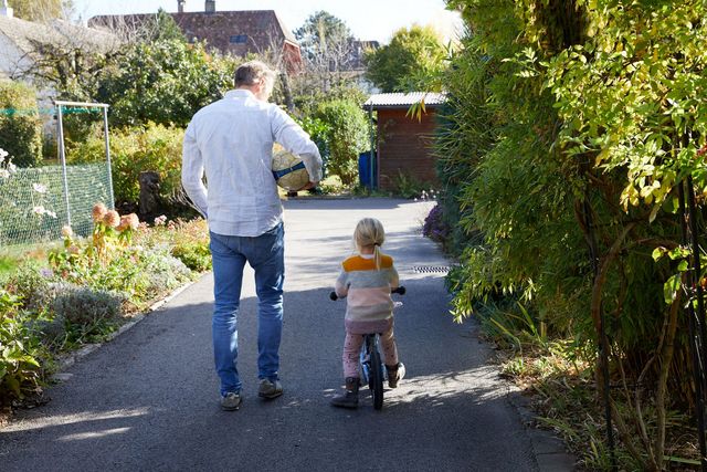 Ein Vater spaziert mit seinem Kind, das auf dem Velo fährt, und trägt dabei einen Ball